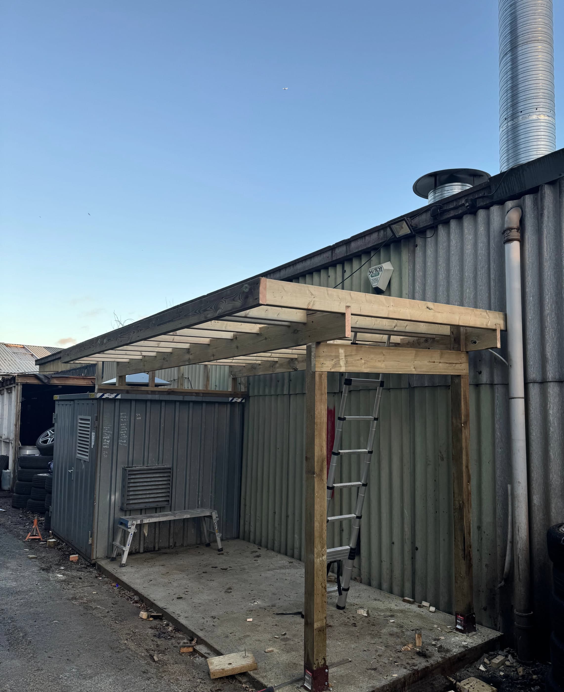 Structural timber lean-to canopy framed against a steel-clad commercial building