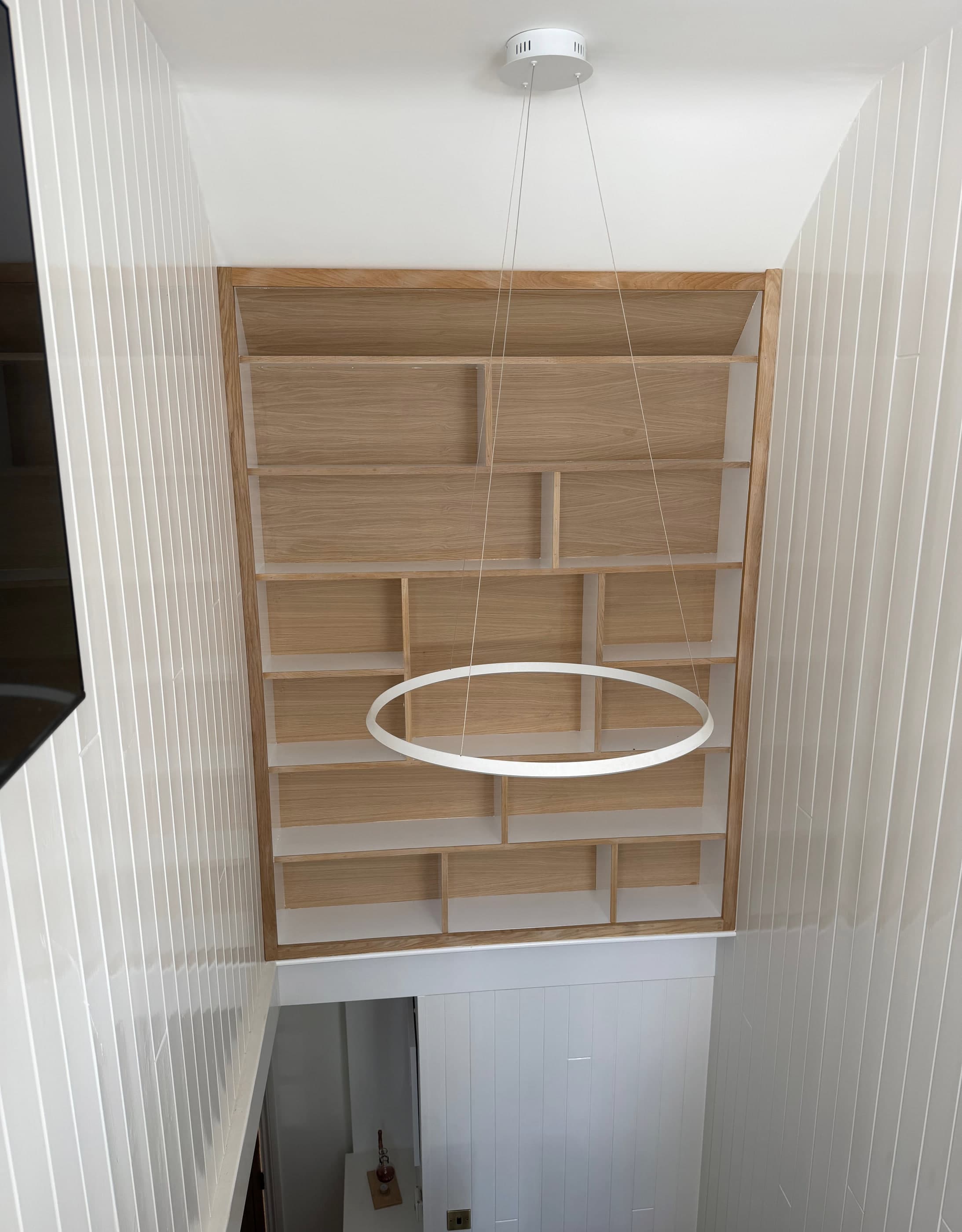 Bespoke oak shelving grid built into a double-height stairwell with white tongue-and-groove panelling and a ring pendant light