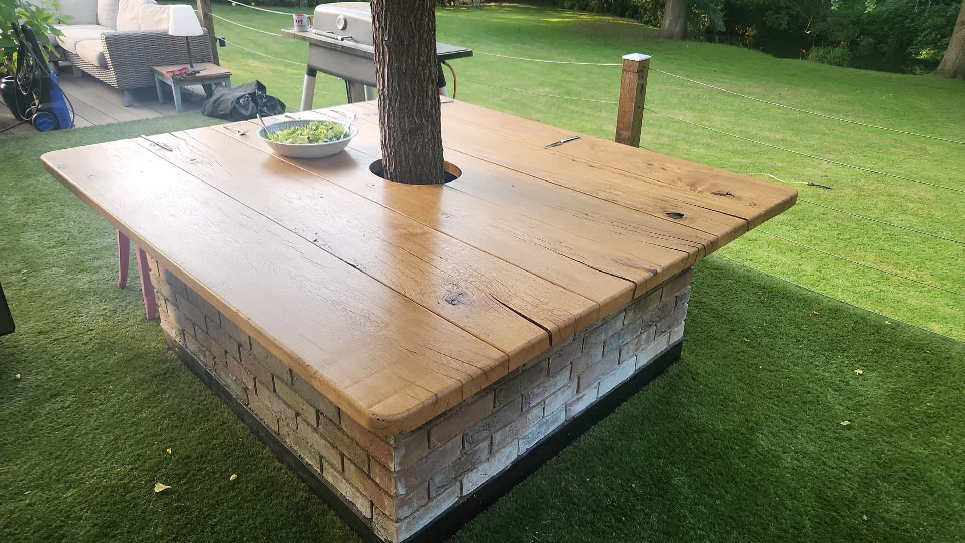 Bespoke solid oak garden table built around a living tree trunk, set on a reclaimed brick plinth on a lawn